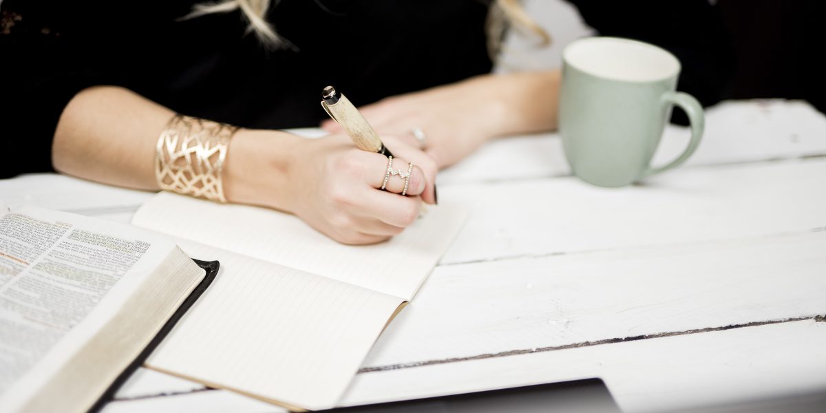 Main écrivant dans un carnet posé sur un livre ouvert, tasse à côté